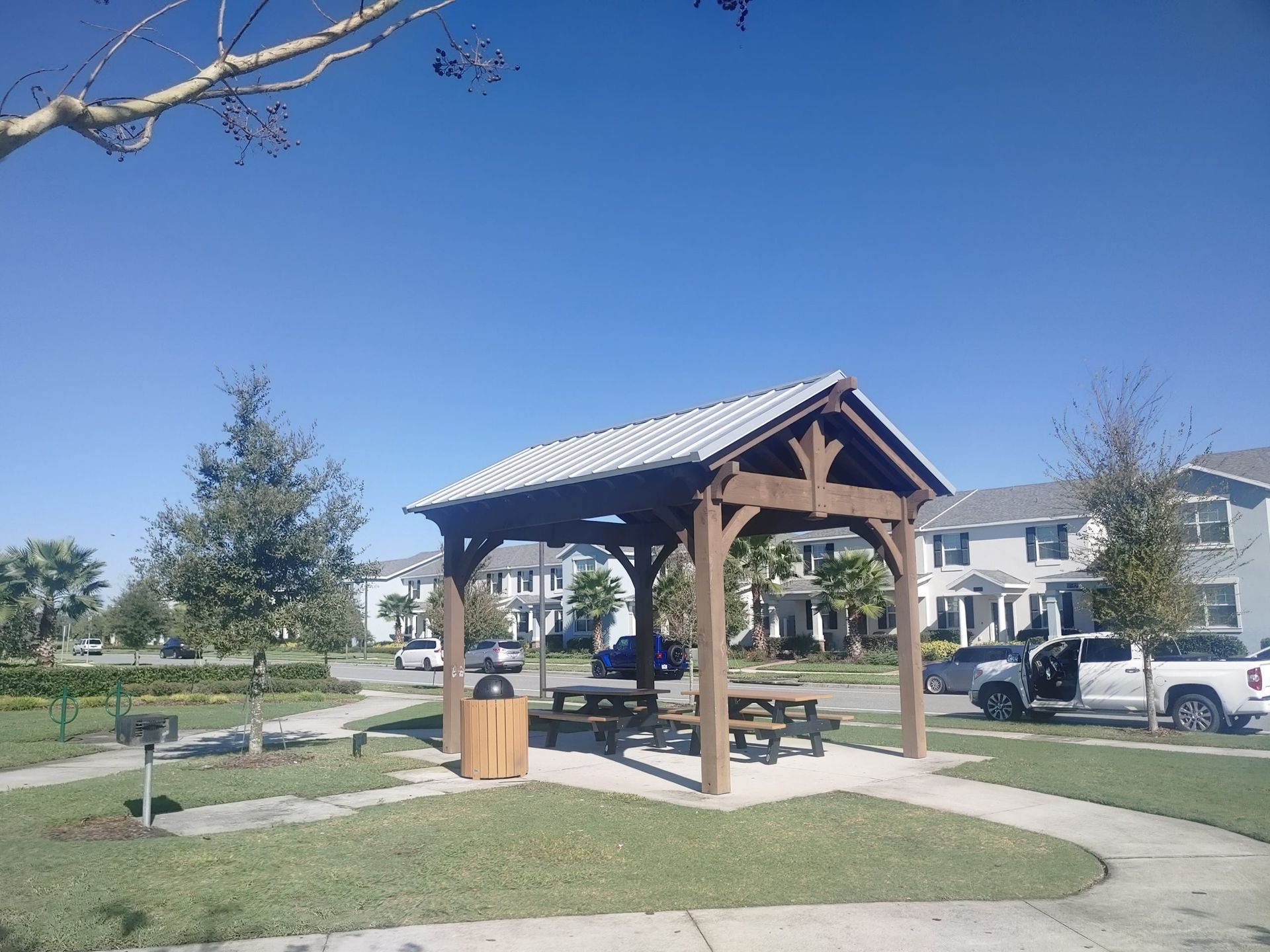 Picnic shelter with tables, grill, and trash can in a park setting with townhouses in the background under a blue sky.