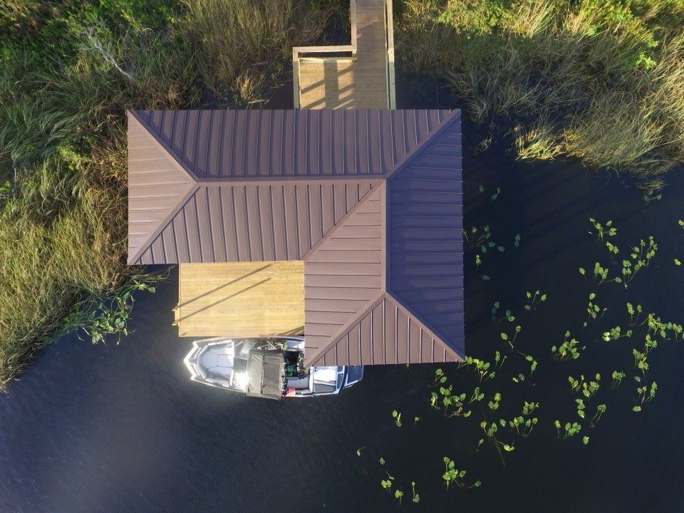 Overhead view: Brown-roofed boathouse over water, boat inside. Green reeds and water lilies surround.