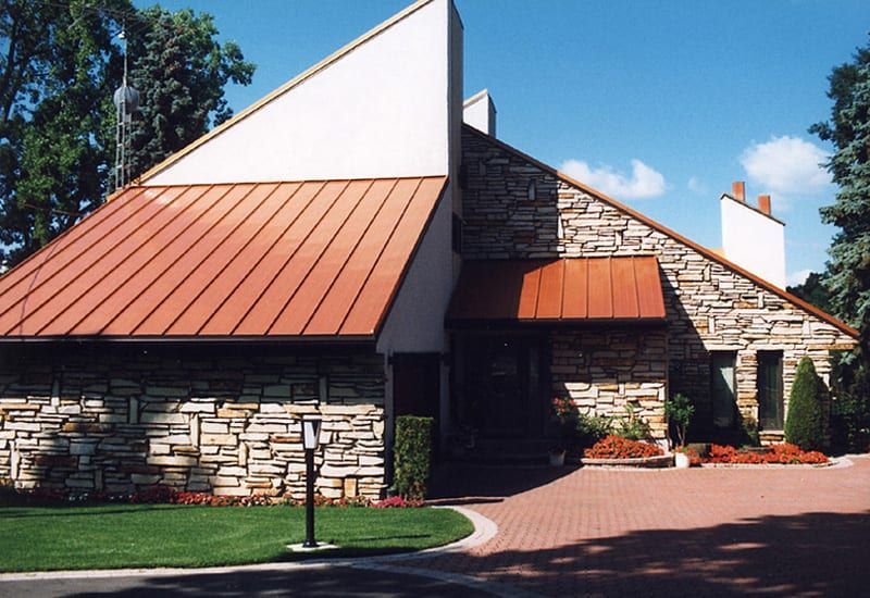 Tan stone house with angled copper roof and brick driveway.