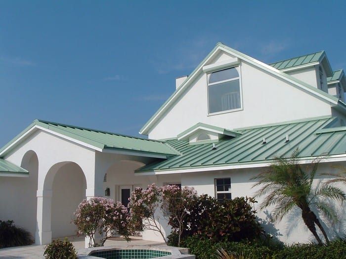 White house with green metal roof and landscaping, set against a clear blue sky.