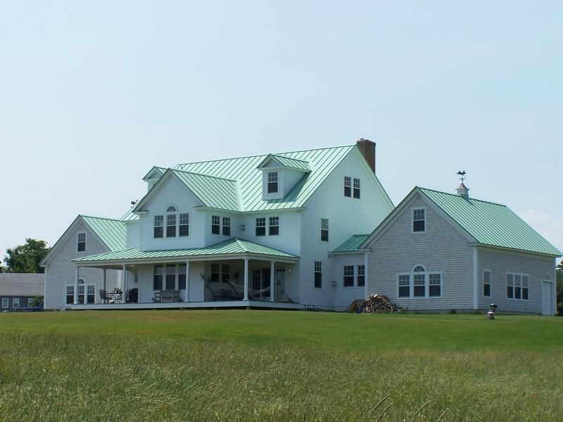 White farmhouse with green roof and porch on a grassy hill under a blue sky.