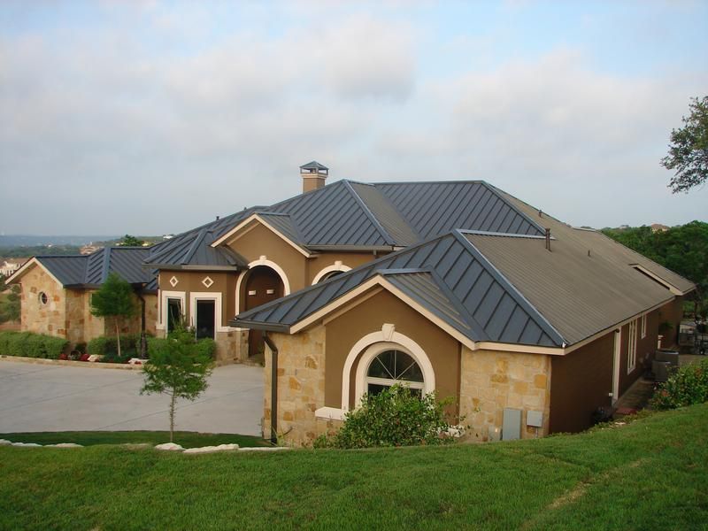 Brown stucco and stone house with a dark blue metal roof, on a grassy hill.