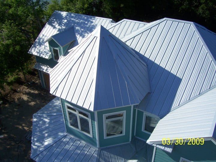 White metal roof on a teal house with multiple angles, surrounded by trees.