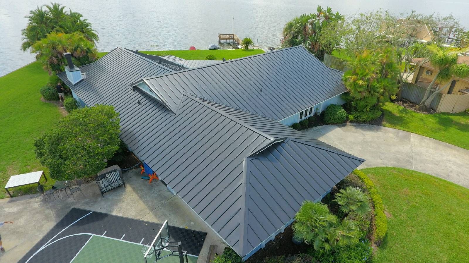 Aerial view of a house with a black roof near water. A basketball court is in the foreground.