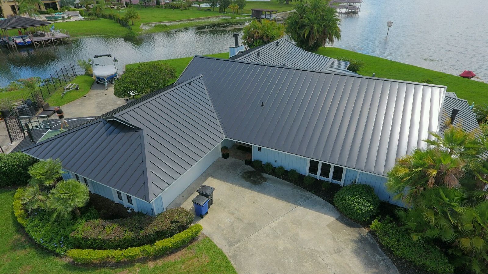 Aerial view of a light blue house with a gray metal roof near a lake, with lush green surroundings.