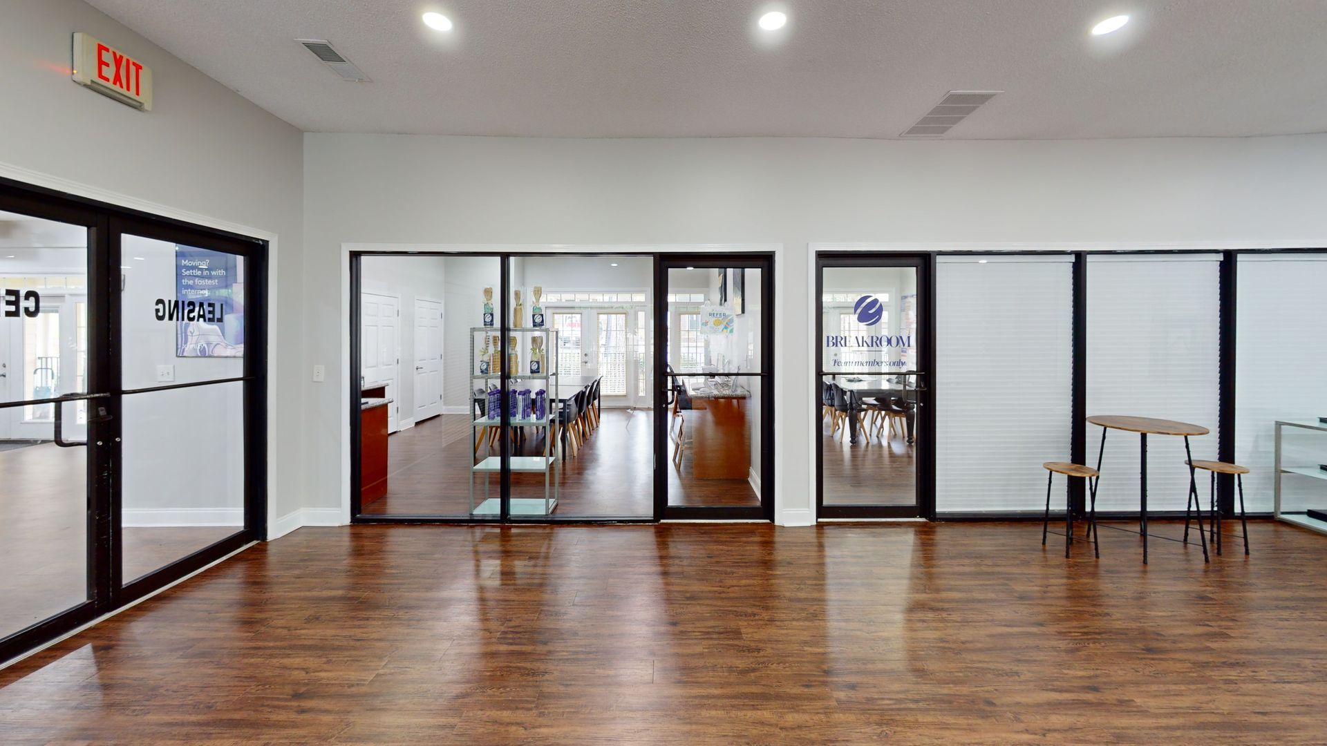Empty modern office with wood floors, glass-walled rooms, and chairs along the right wall