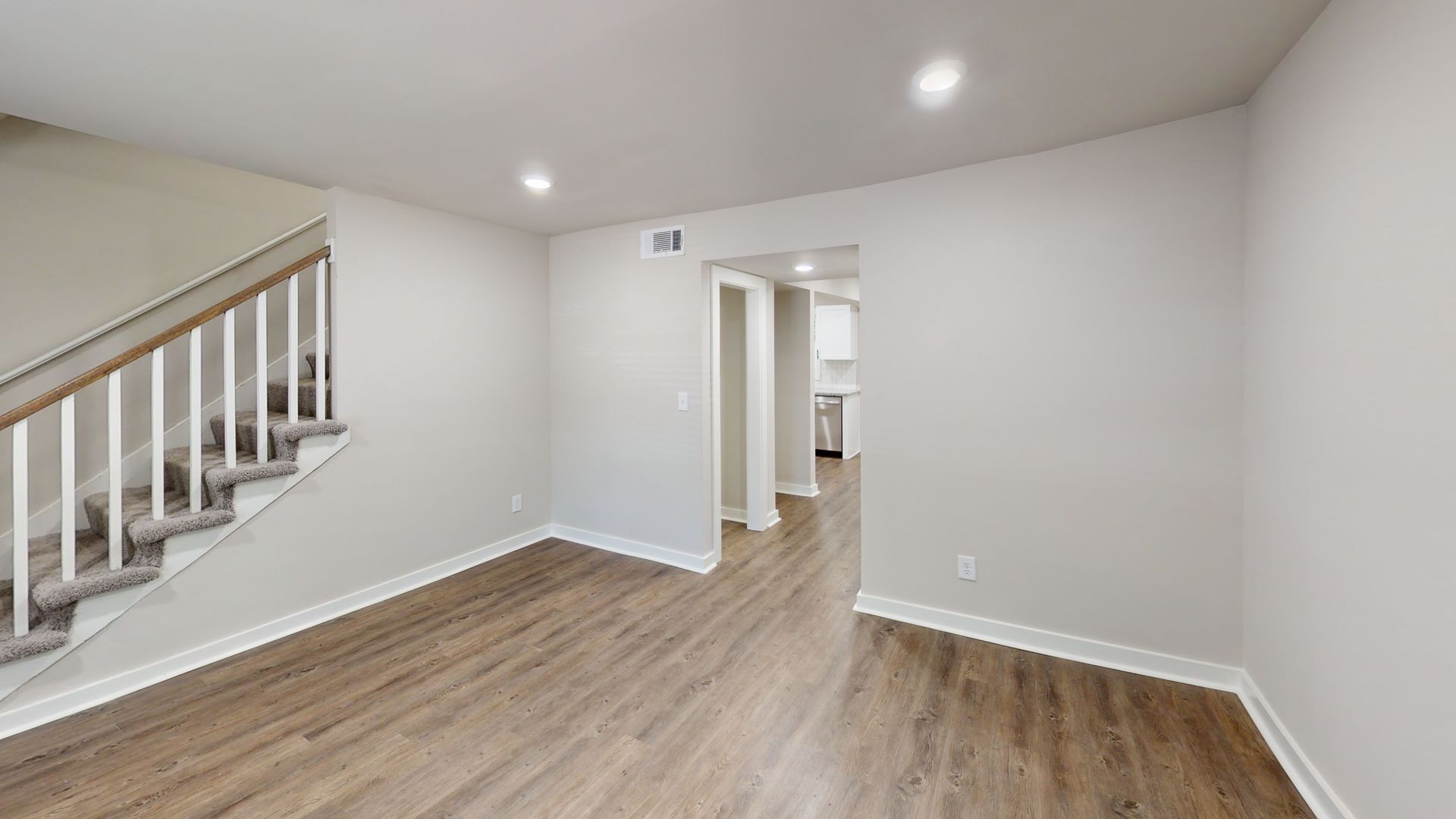 Empty modern room with gray walls, wood floors, recessed lights, and a staircase on the left