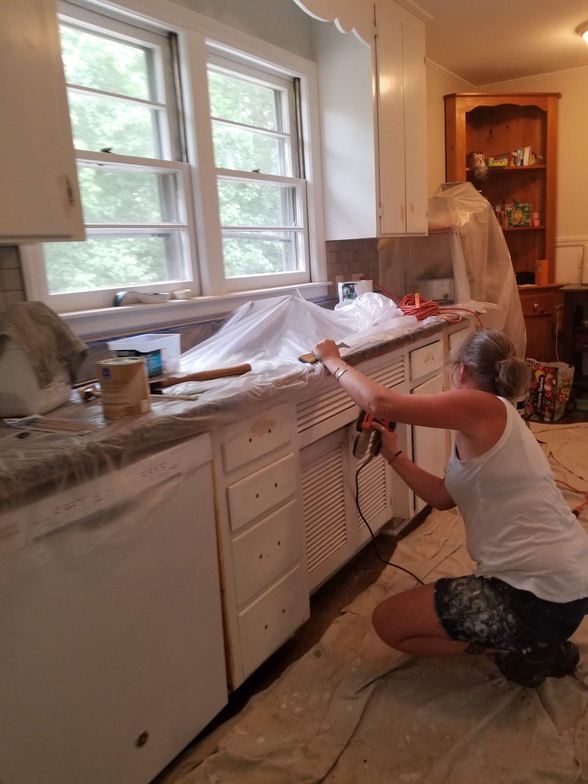 A woman is kneeling down in a kitchen painting cabinets.