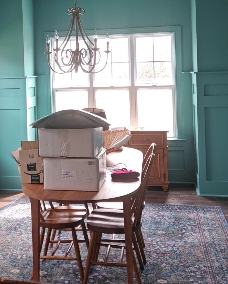 Dining room with teal walls, a wooden table with boxes, chairs, rug, and chandelier.