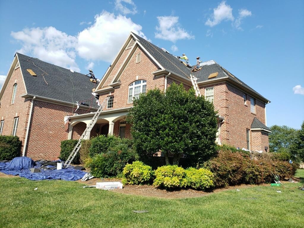 Brick suburban house with a gabled roof, green lawn, shrubs, and blue tarp under a bright sky