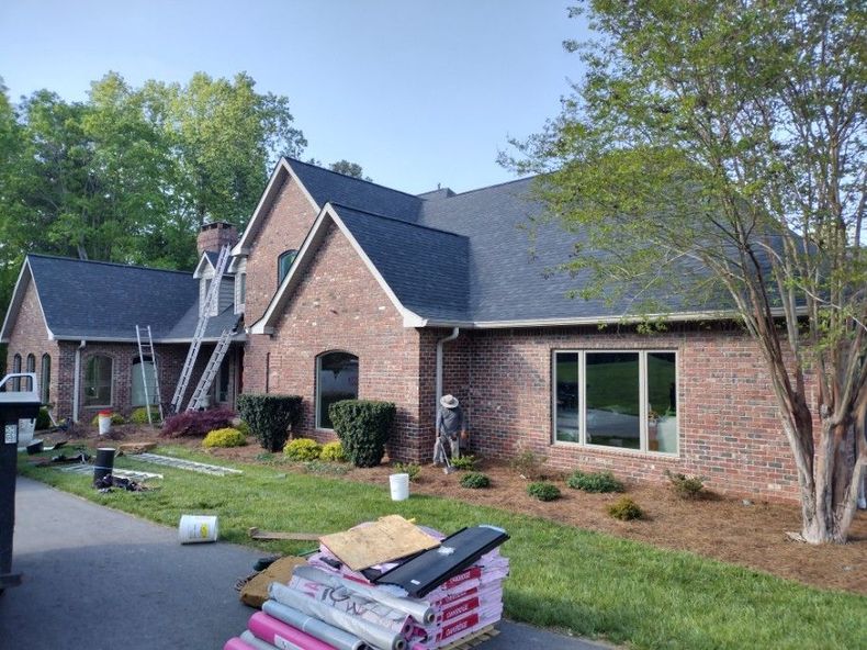Brick suburban house with landscaped yard and stacked roofing materials in the foreground