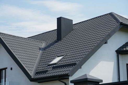 Modern house roof with dark gray tiles, skylight, and chimney against a blue sky