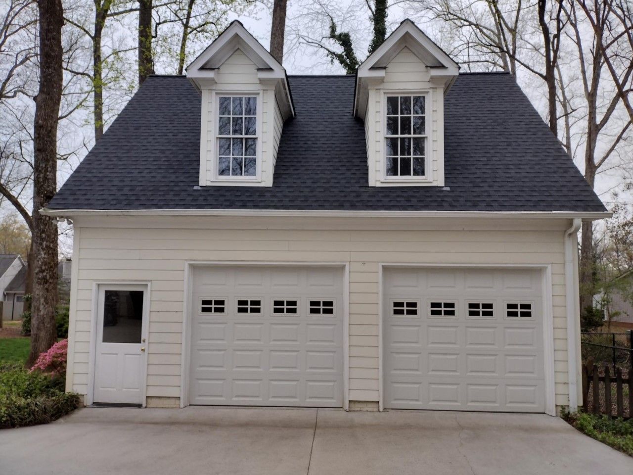 White two-car garage with dormer windows and a side door in a wooded residential setting
