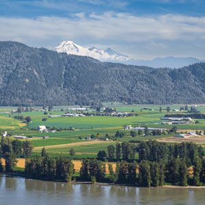 Scenic view: river in foreground, farmland, forested hills, and snowy mountain peak under blue sky.