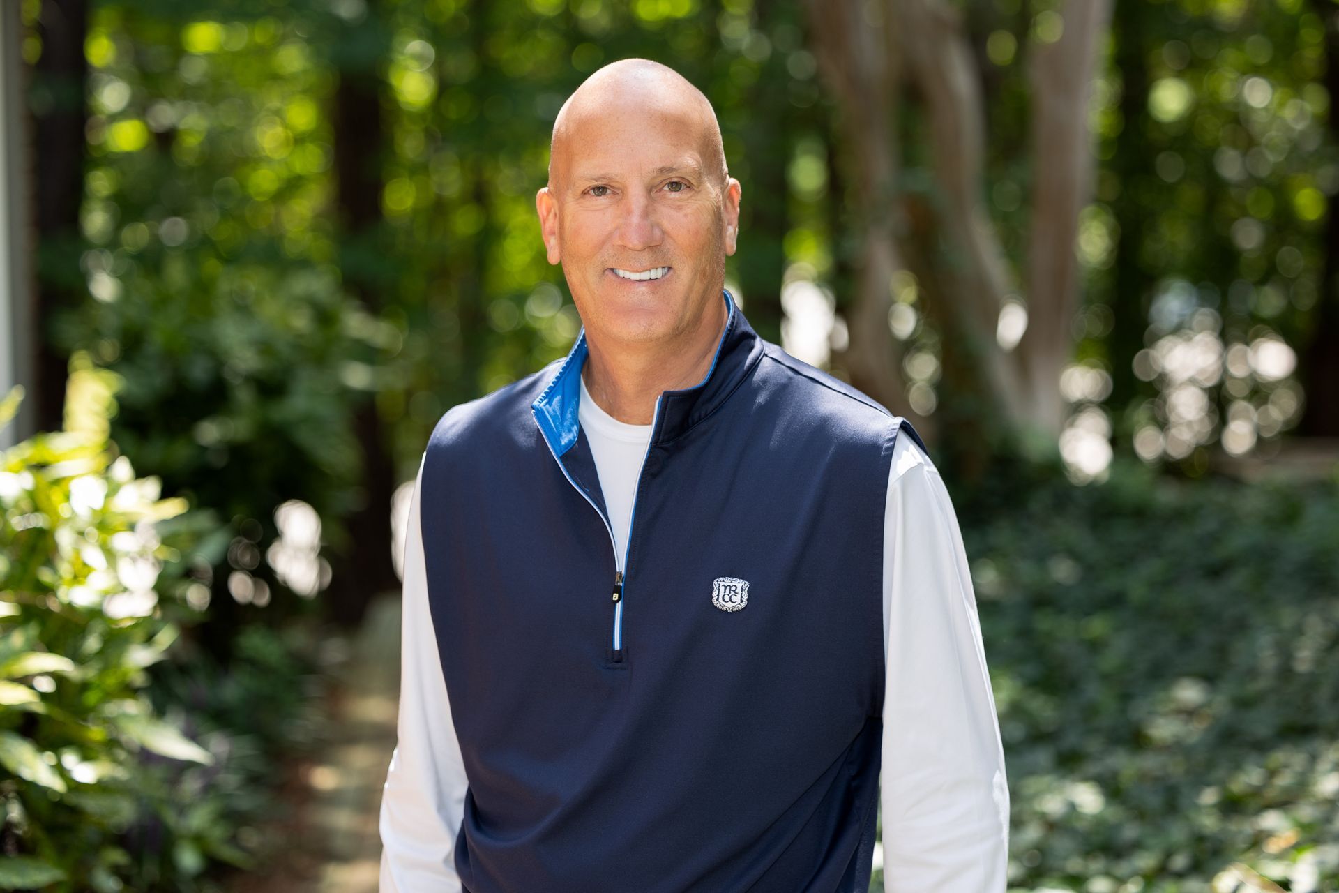 A bald man wearing a blue vest and a white shirt is standing in the woods.