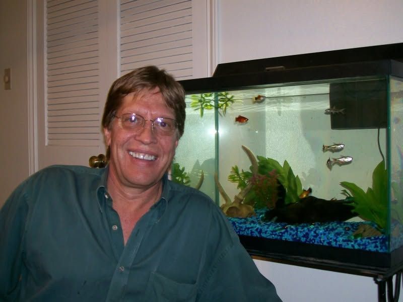 Smiling man in a green shirt beside a lit aquarium with fish and plants