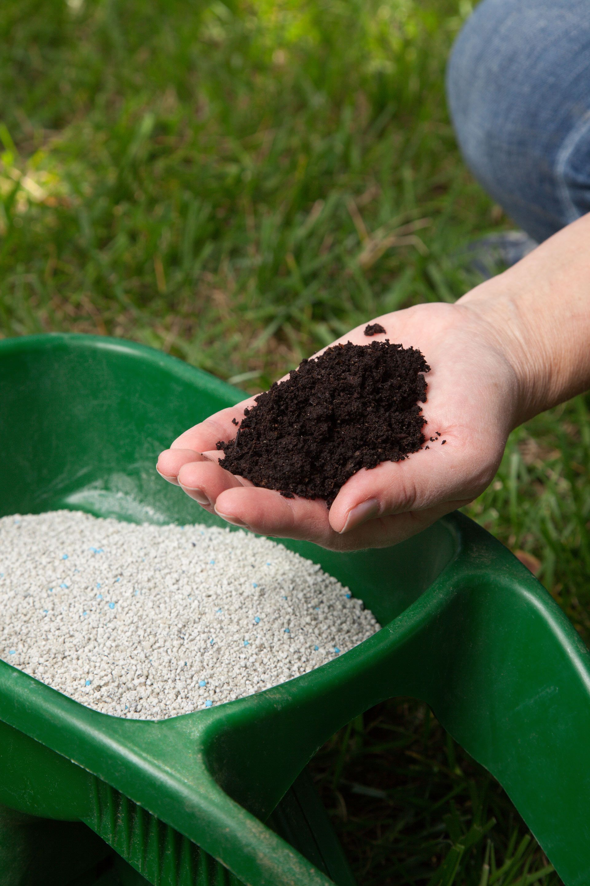 A hand holds a scoop of dark soil over a green garden cart filled with granular white fertilizer.