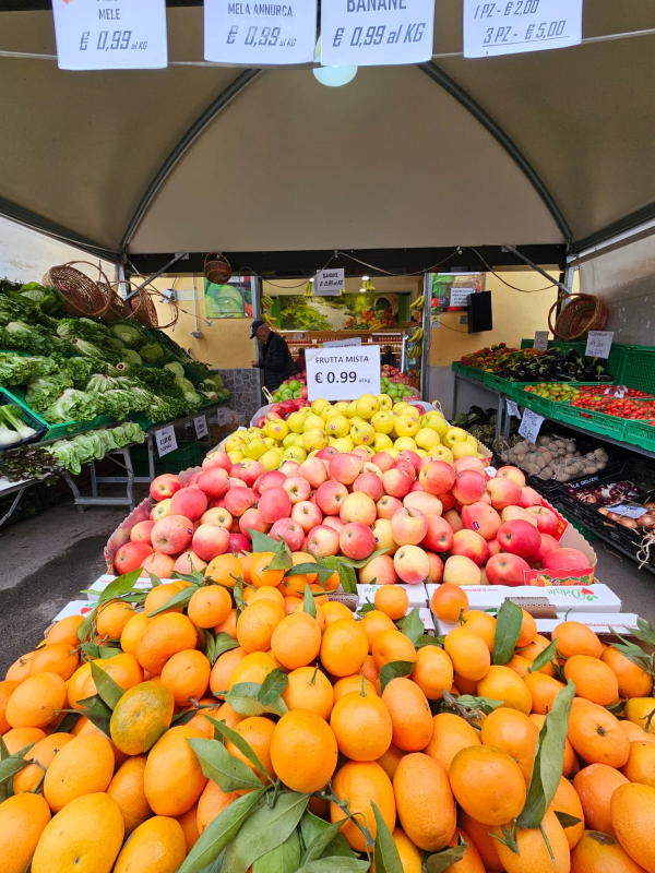Bancarella di frutta e verdura con arance, mele e altri prodotti, una persona sullo sfondo.
