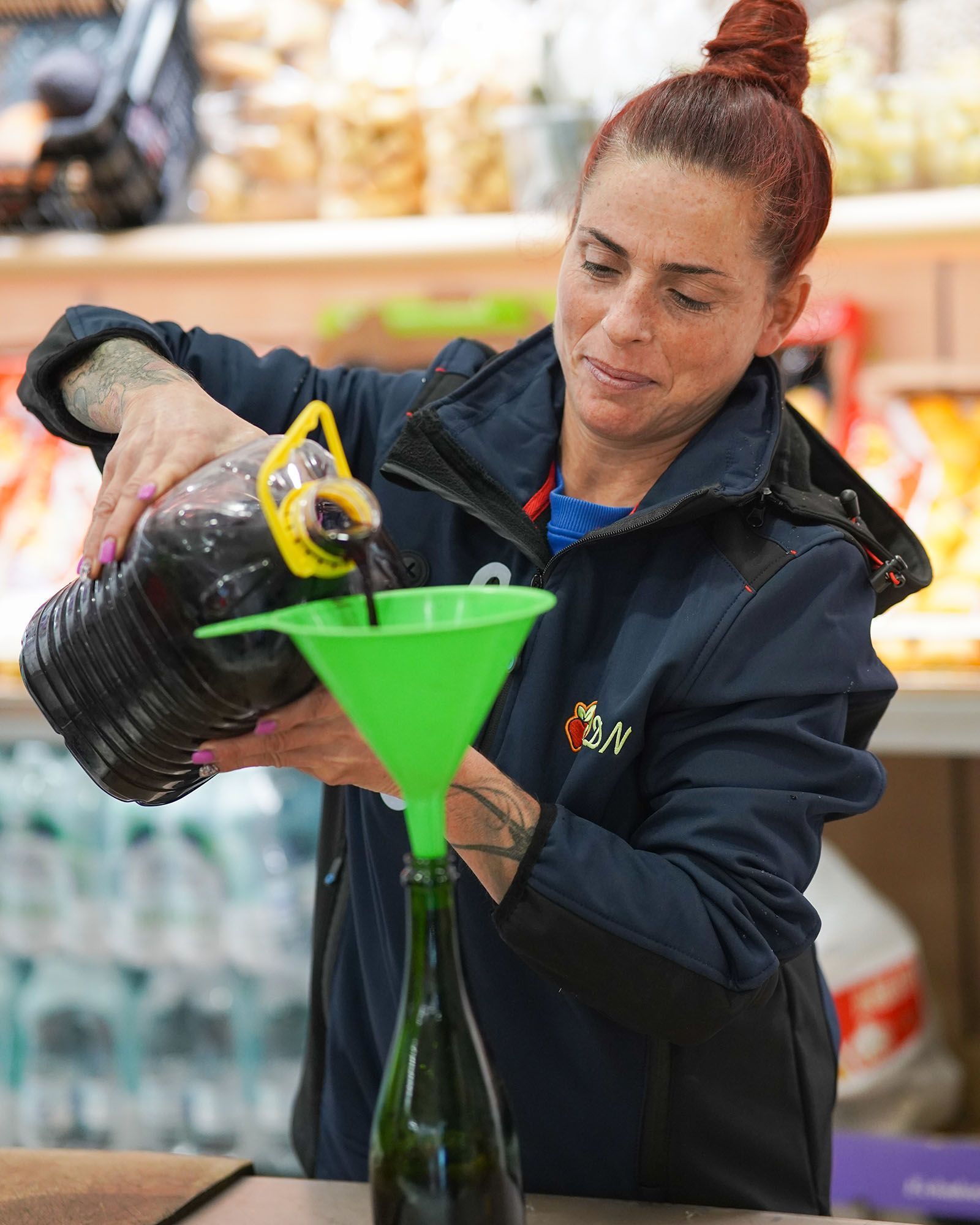 Donna che versa un liquido scuro da un grande contenitore in una bottiglia usando un imbuto.