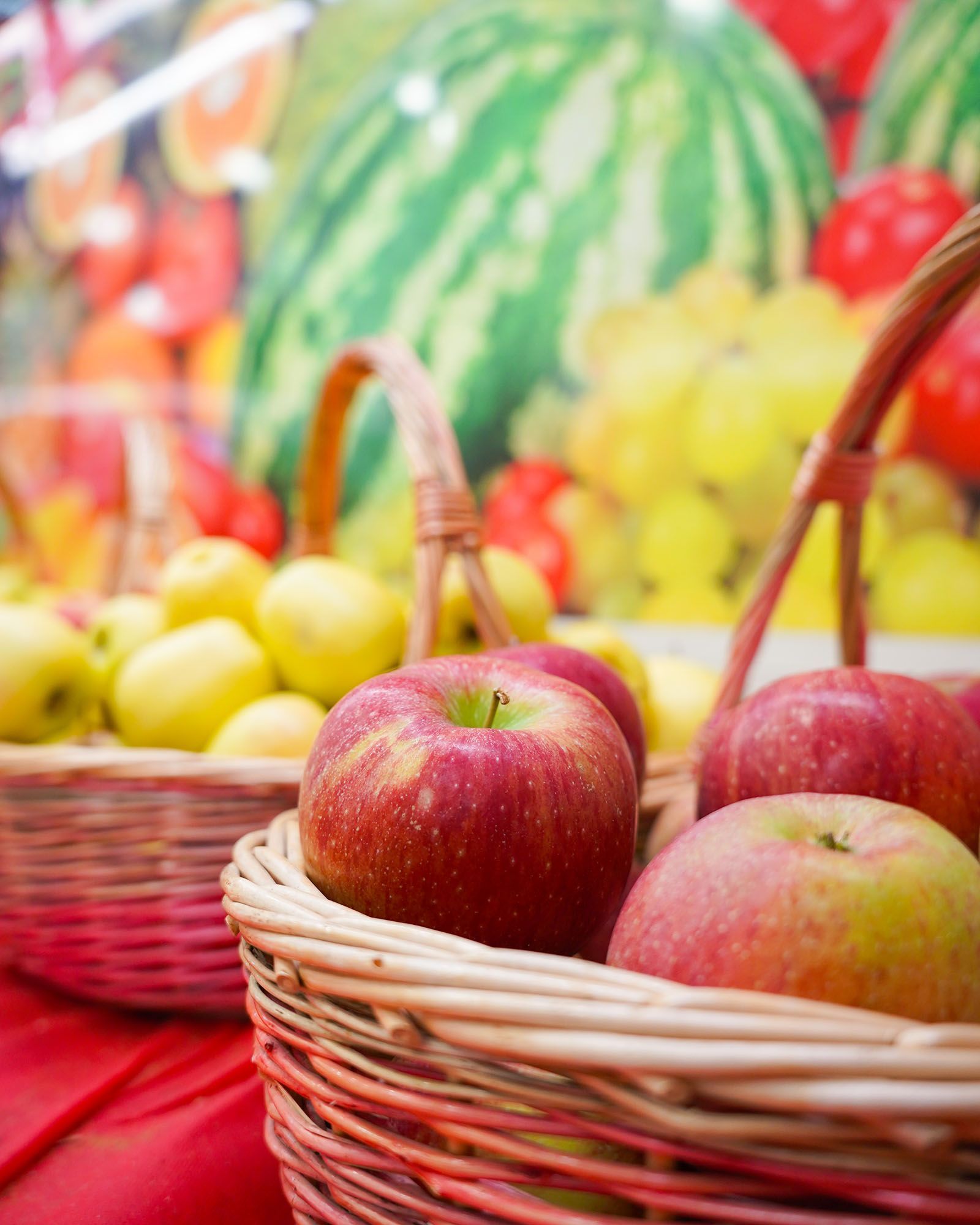 Cestini di vimini pieni di mele rosse e gialle, disposti davanti a una colorata esposizione di frutta.