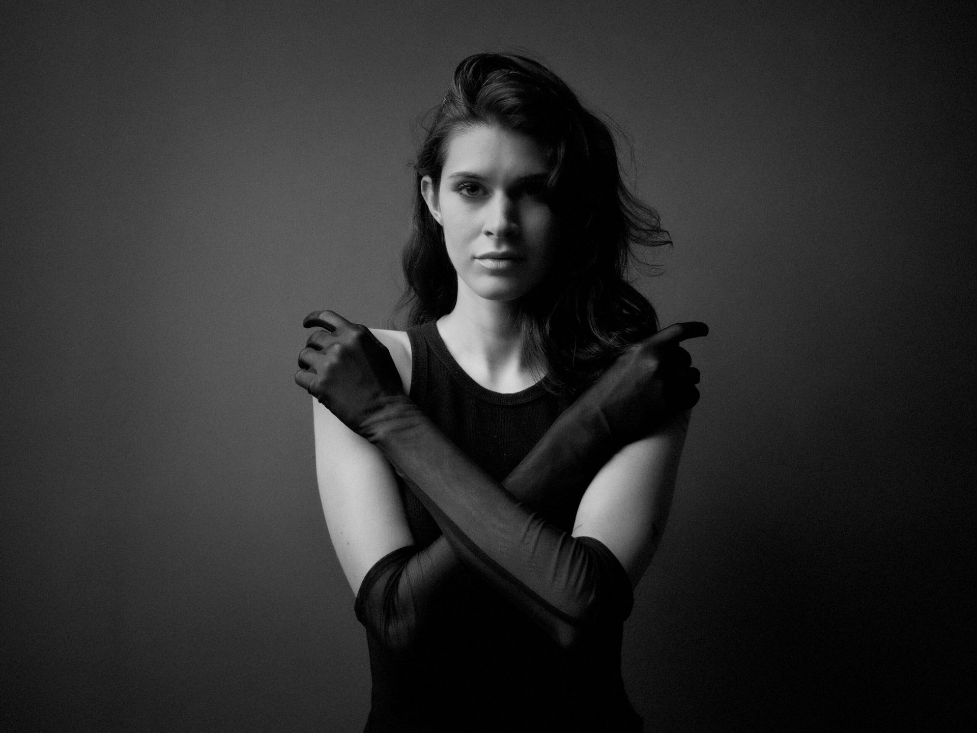 Une femme en robe noire et gants croise les bras devant un fond sombre, le regard droit devant elle.
