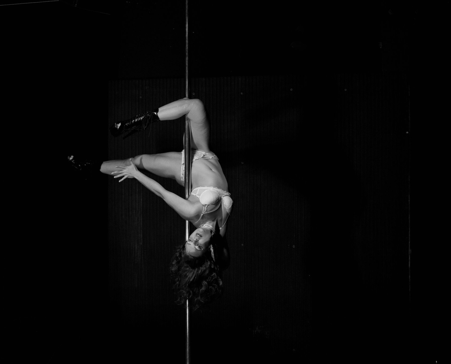 Femme la tête en bas sur une barre, vêtue de lingerie et de bottes, dans un décor sombre.