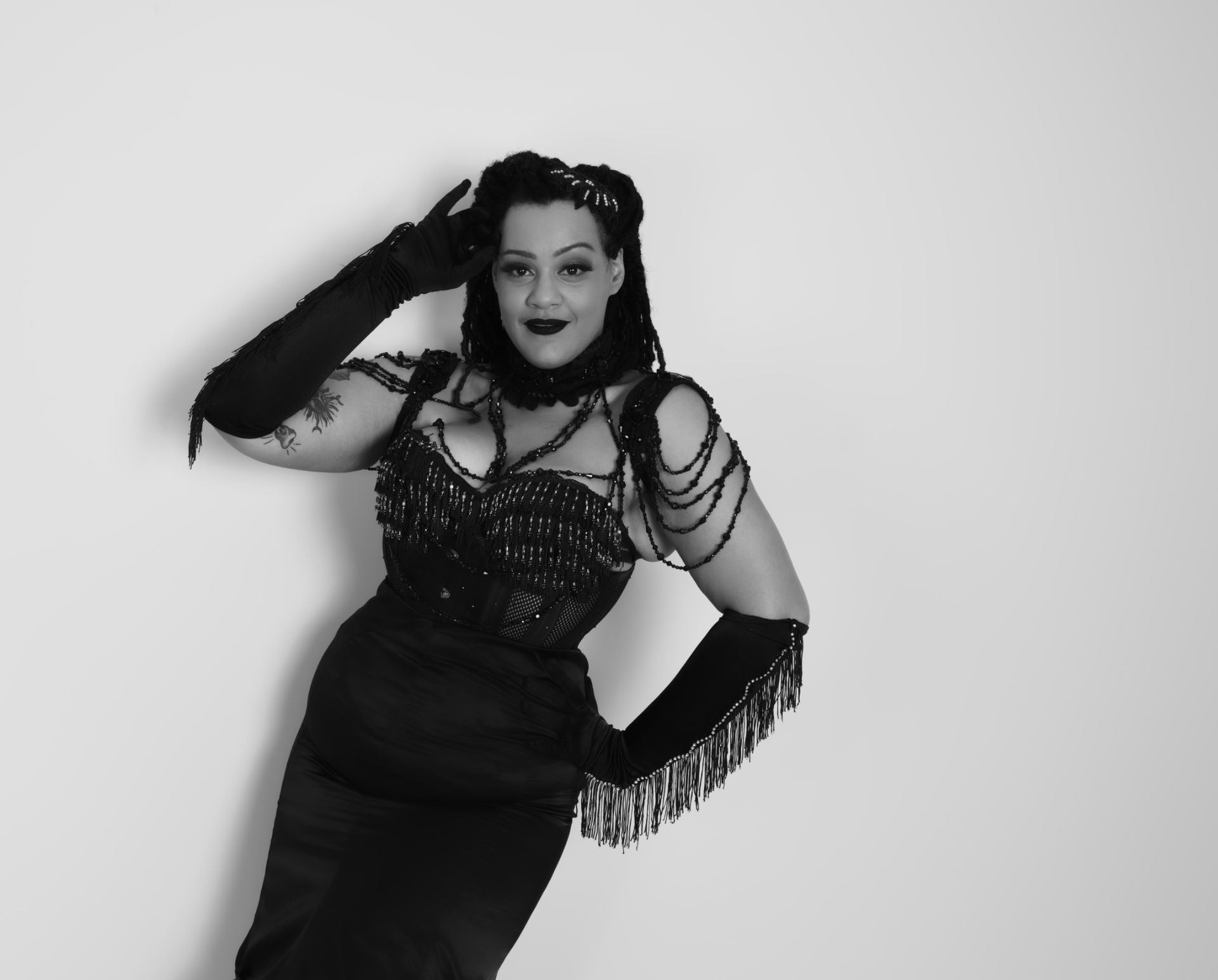 Femme en robe noire, gants et accessoires perlés, posant devant un mur blanc.