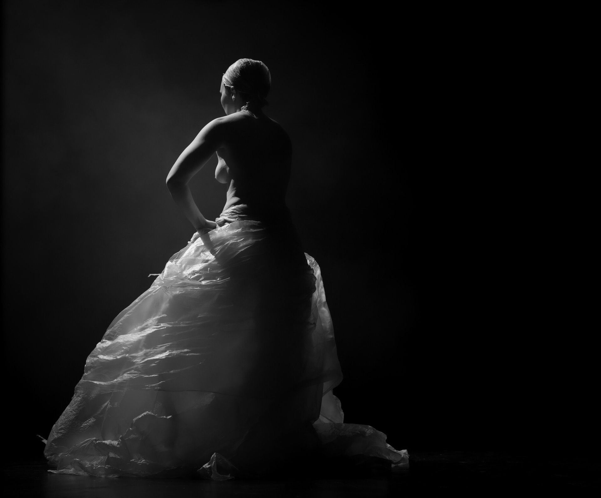 Femme en robe transparente et drapée, à contre-jour sur fond sombre, posant la main sur la hanche.