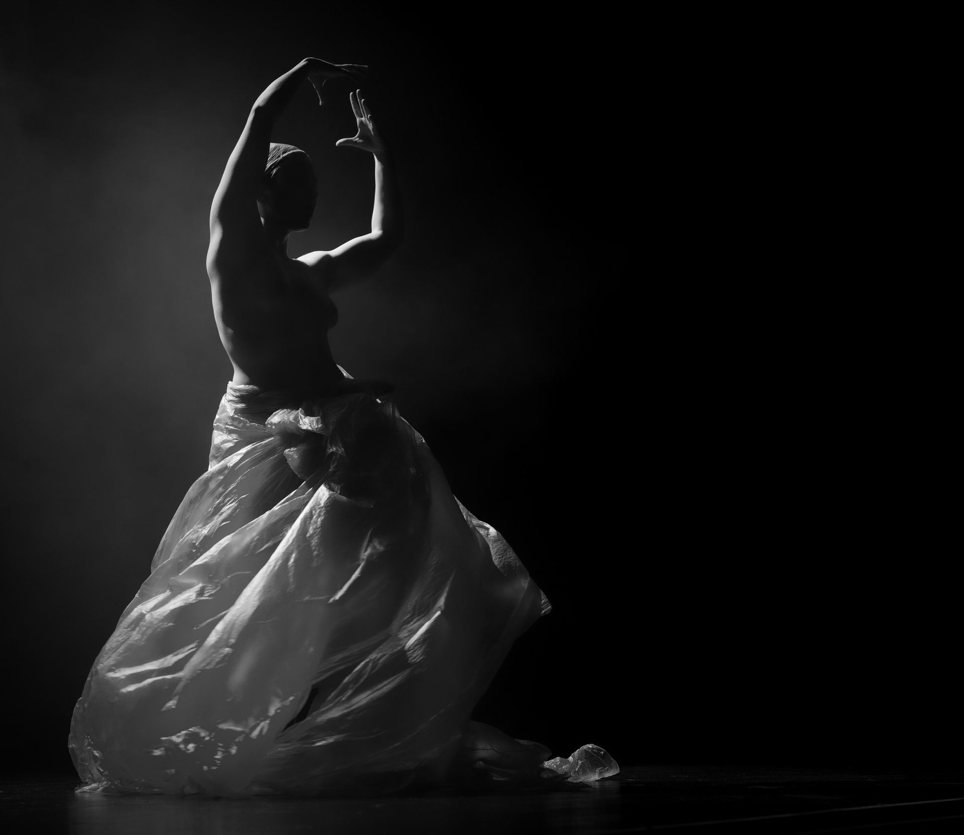 Une personne danse, les bras levés, enveloppée dans un tissu translucide sur un fond sombre.