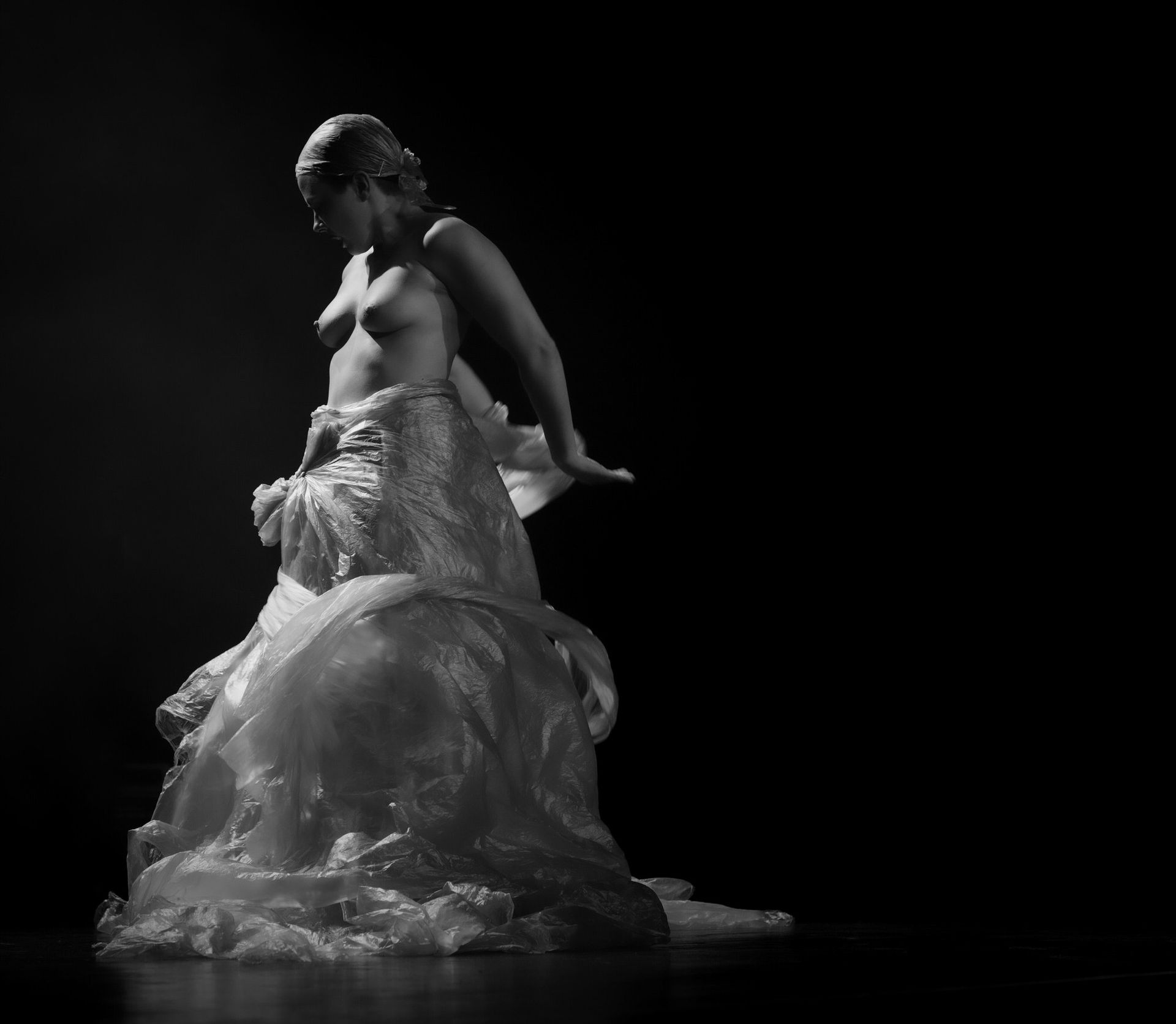Femme vêtue d'un tissu translucide sur fond sombre ; bras tendus, regard baissé.