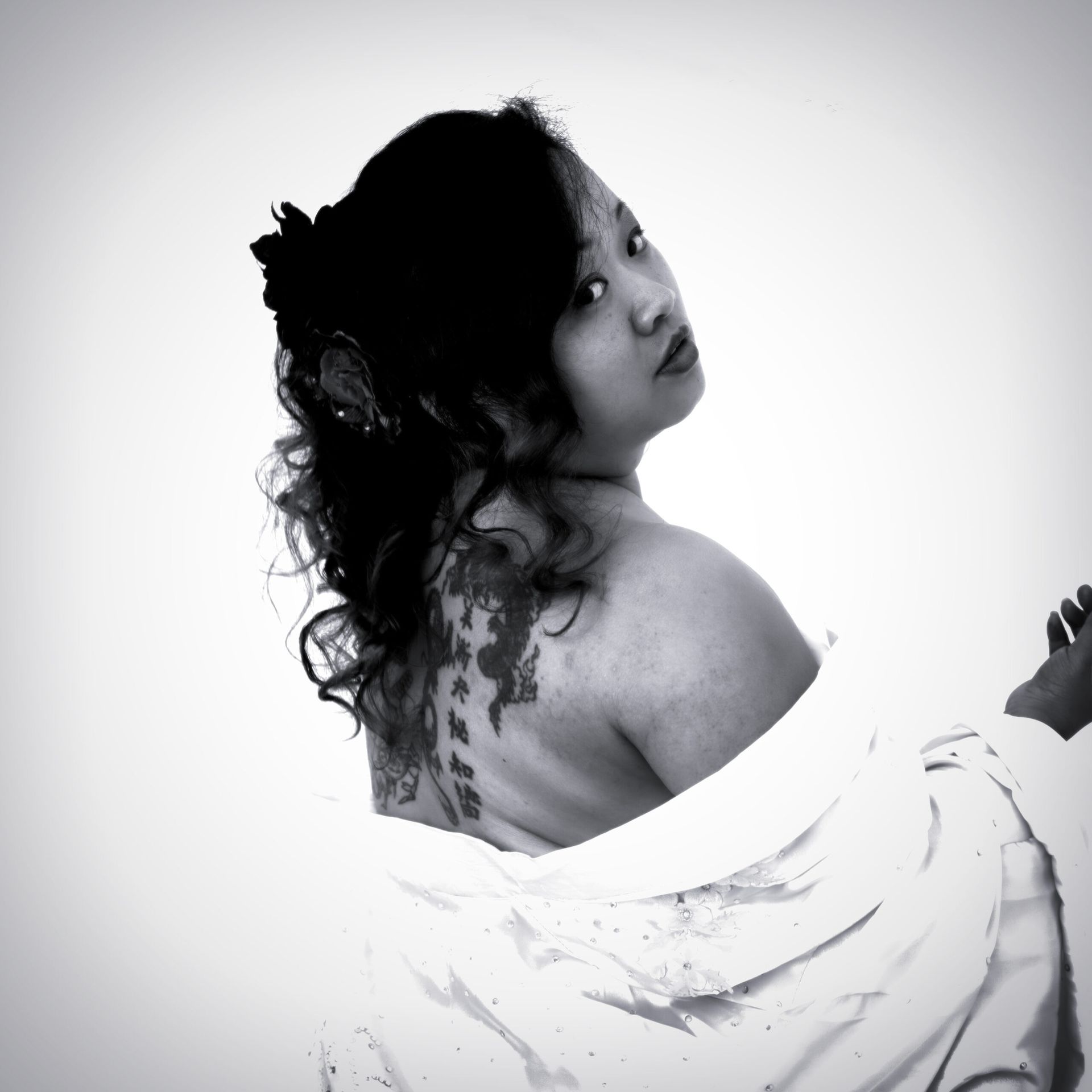 Femme tatouée dans le dos, vêtue d'une robe de chambre, regardant par-dessus son épaule. Noir et blanc.