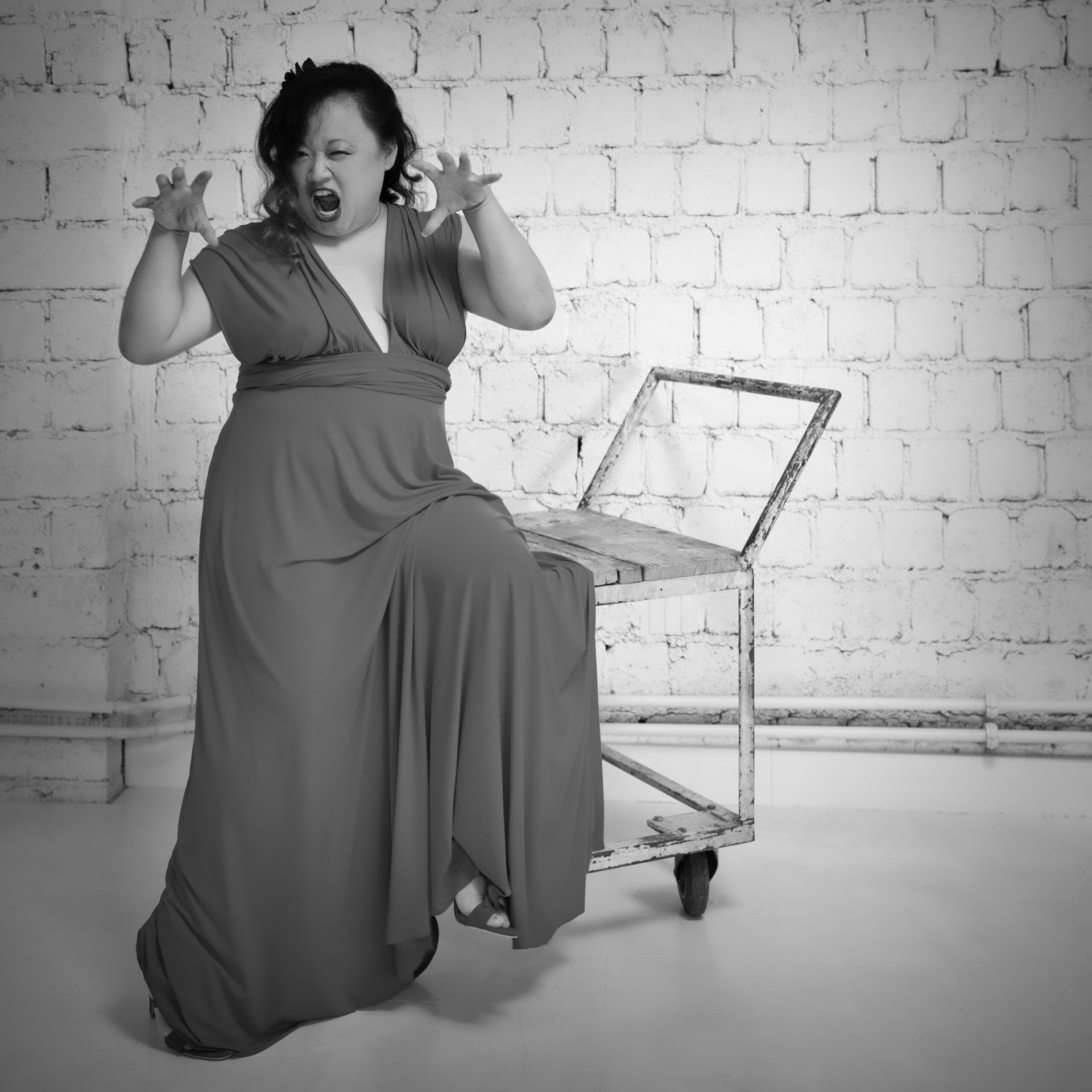 Une femme en robe rouge, les yeux écarquillés, les bras levés, s'appuie sur un chariot métallique contre un mur de briques blanches.