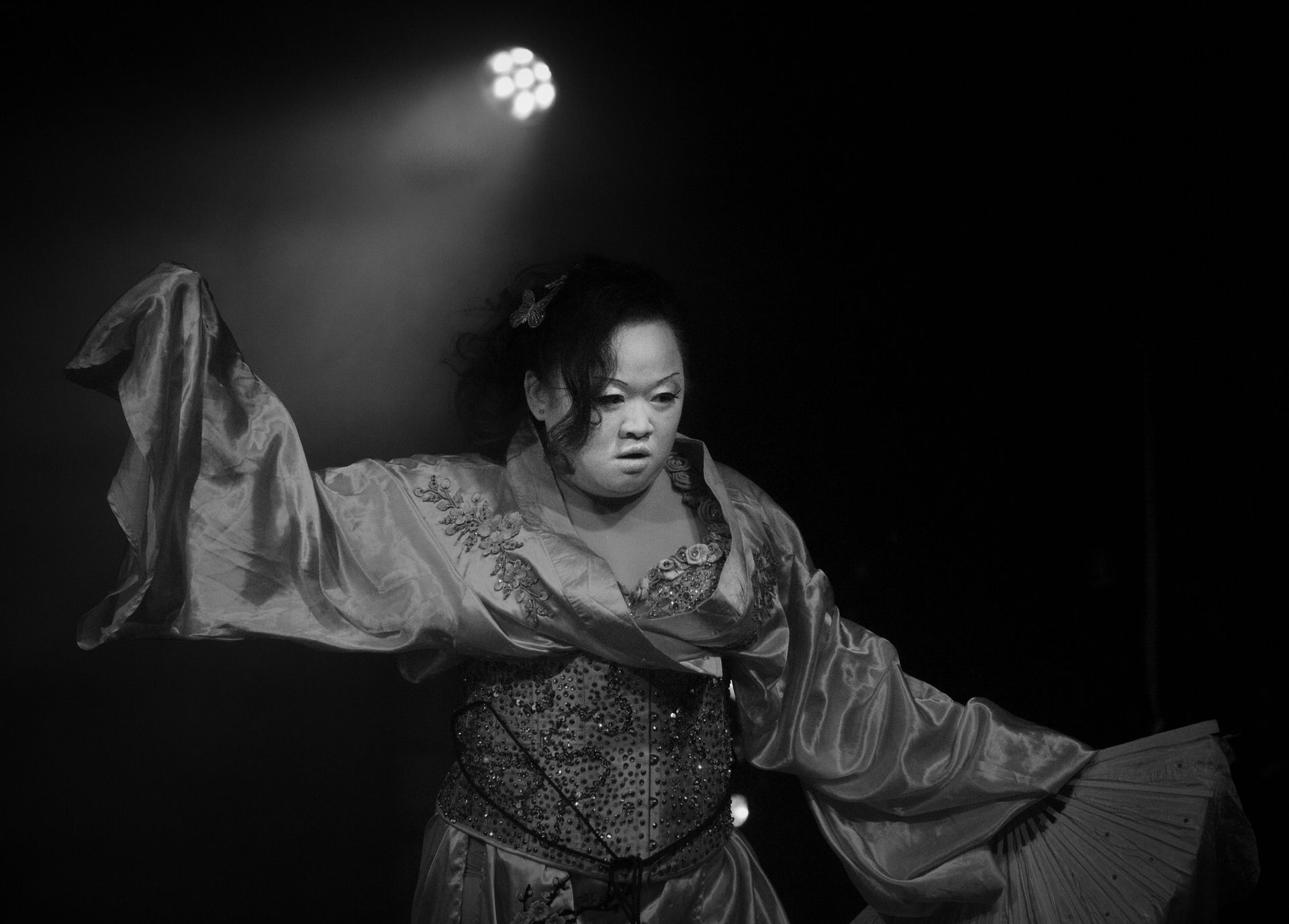 Une artiste en costume élaboré aux manches flottantes, les bras tendus sous un projecteur, en noir et blanc.