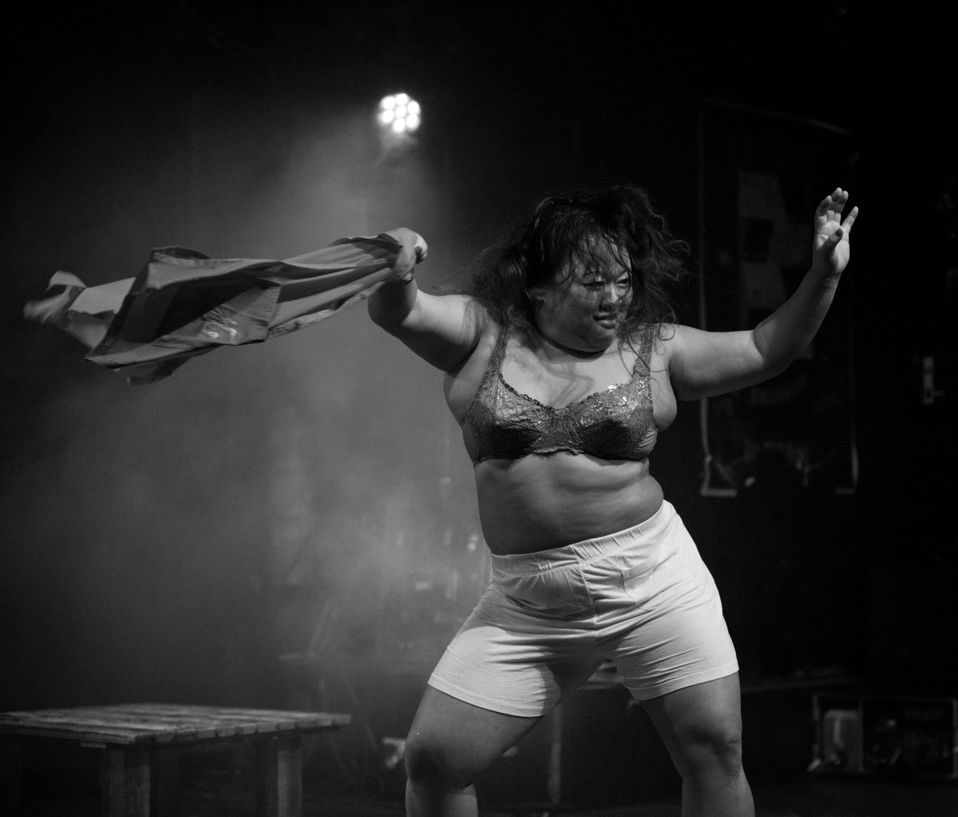 Une personne en lingerie sur scène, tenant un vêtement, éclairée par un projecteur, expressive.