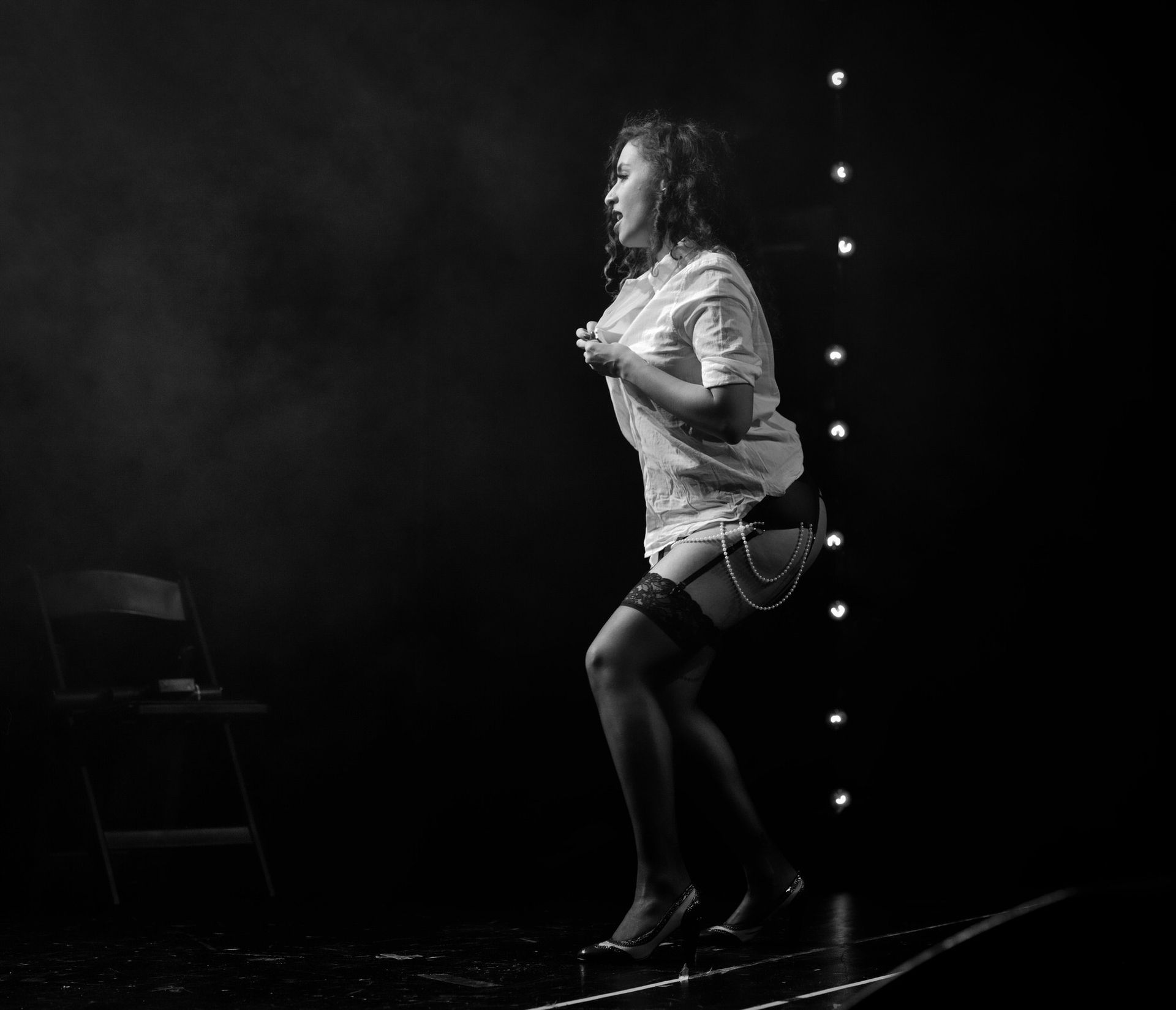 Femme en lingerie et chemise oversize sur scène, dansant à côté d'une chaise.