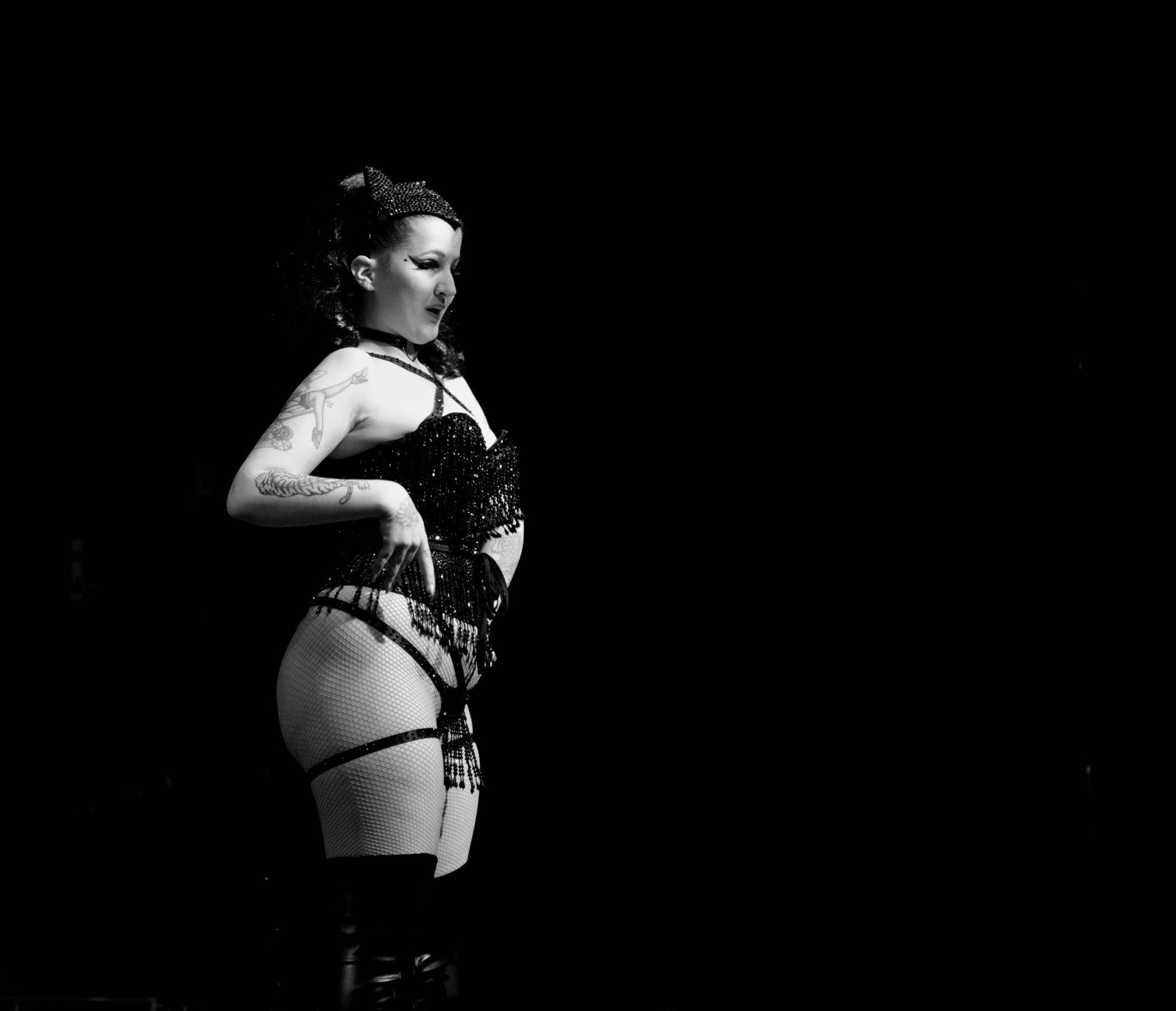 Femme en lingerie noire, tatouages, main sur la hanche, regardant à droite, sur fond noir.