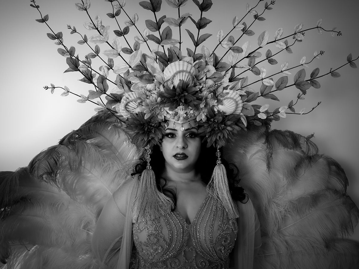 Femme coiffée d'une parure florale élaborée, vêtue d'une robe ornée, se tenant devant un décor d'ailes.