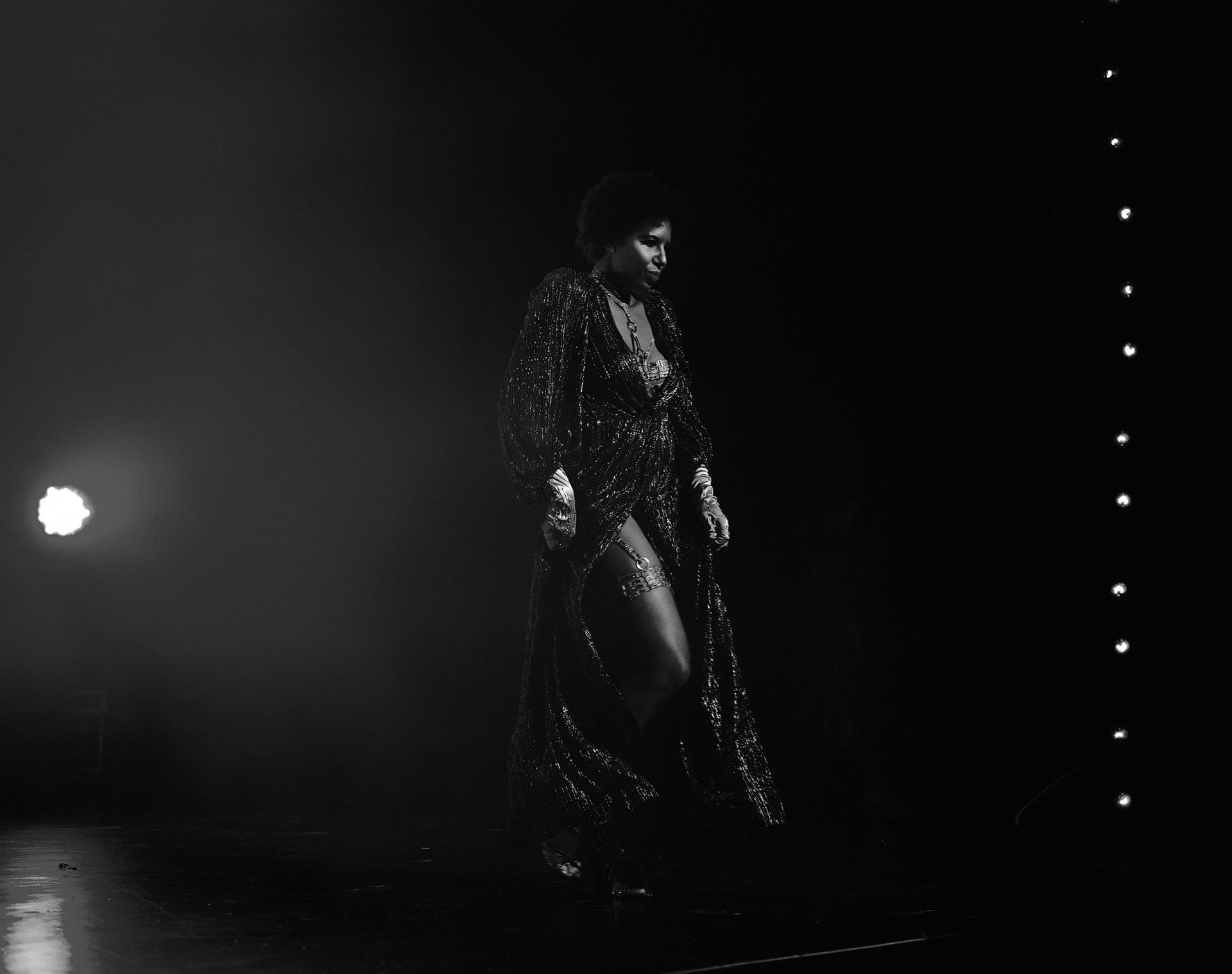 Femme en tenue scintillante sur scène, marchant vers la droite, sous les projecteurs.