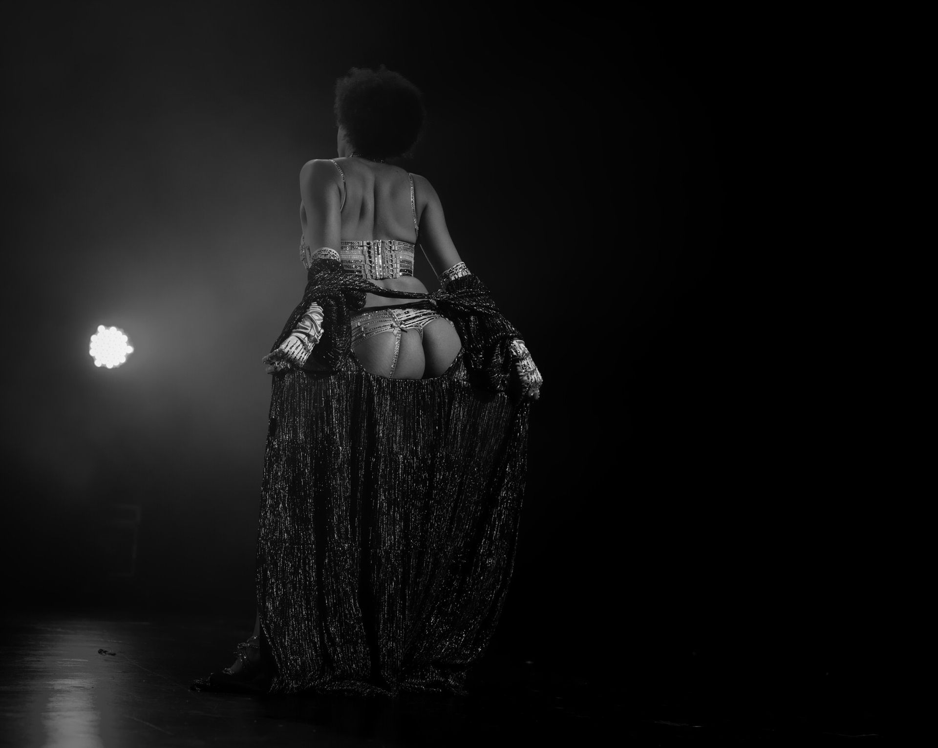 Vue de dos d'une personne en costume orné et scintillant dansant sur une scène sombre éclairée par un projecteur.