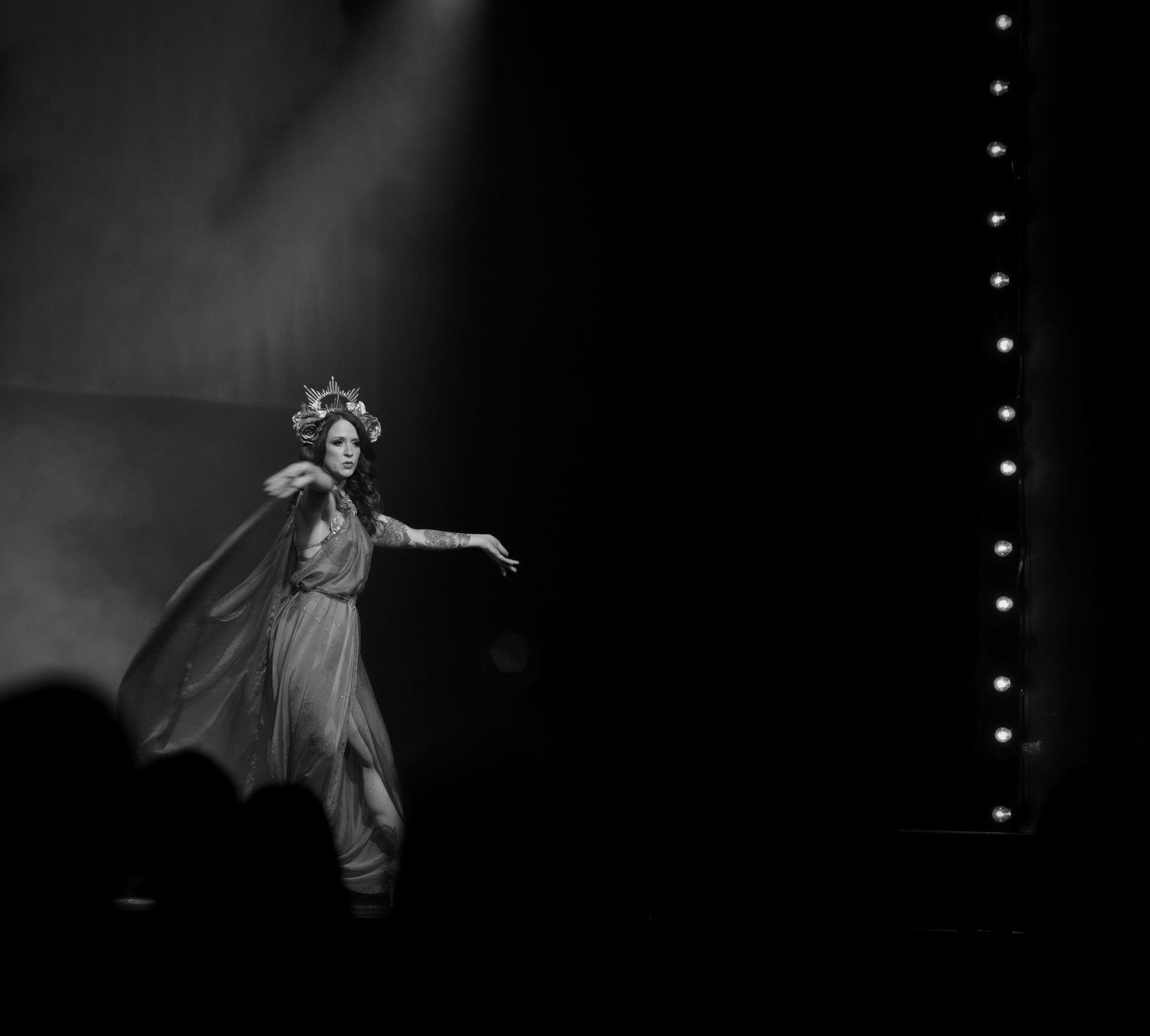 Femme en robe fluide et couronne sur scène, le bras tendu. Projecteur sur elle.