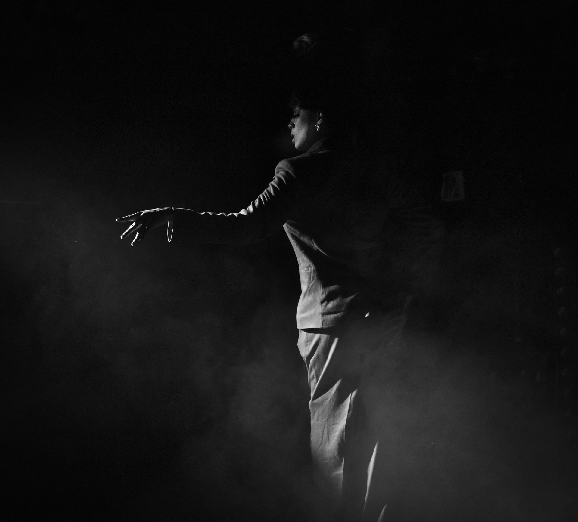 Personne, bras tendu, dans une pièce faiblement éclairée, peut-être en train de danser ; atmosphère enfumée.