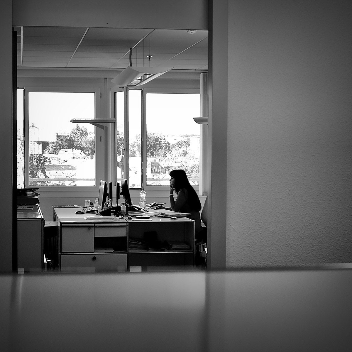 Une personne est assise à un bureau dans un bureau, face à des fenêtres et des ordinateurs. Noir et blanc.