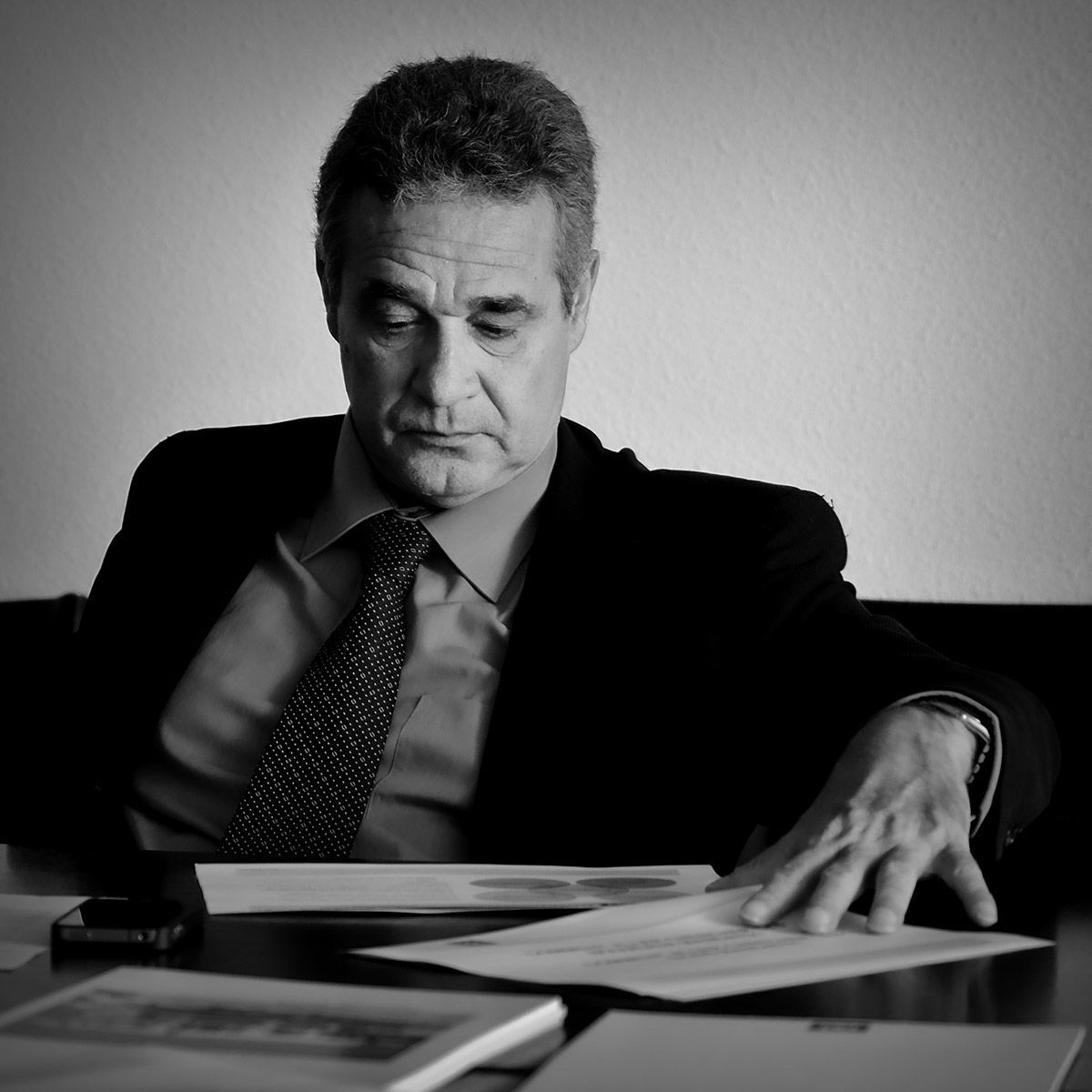 Un homme en costume consulte des papiers posés sur une table, le bras appuyé dessus. Noir et blanc.