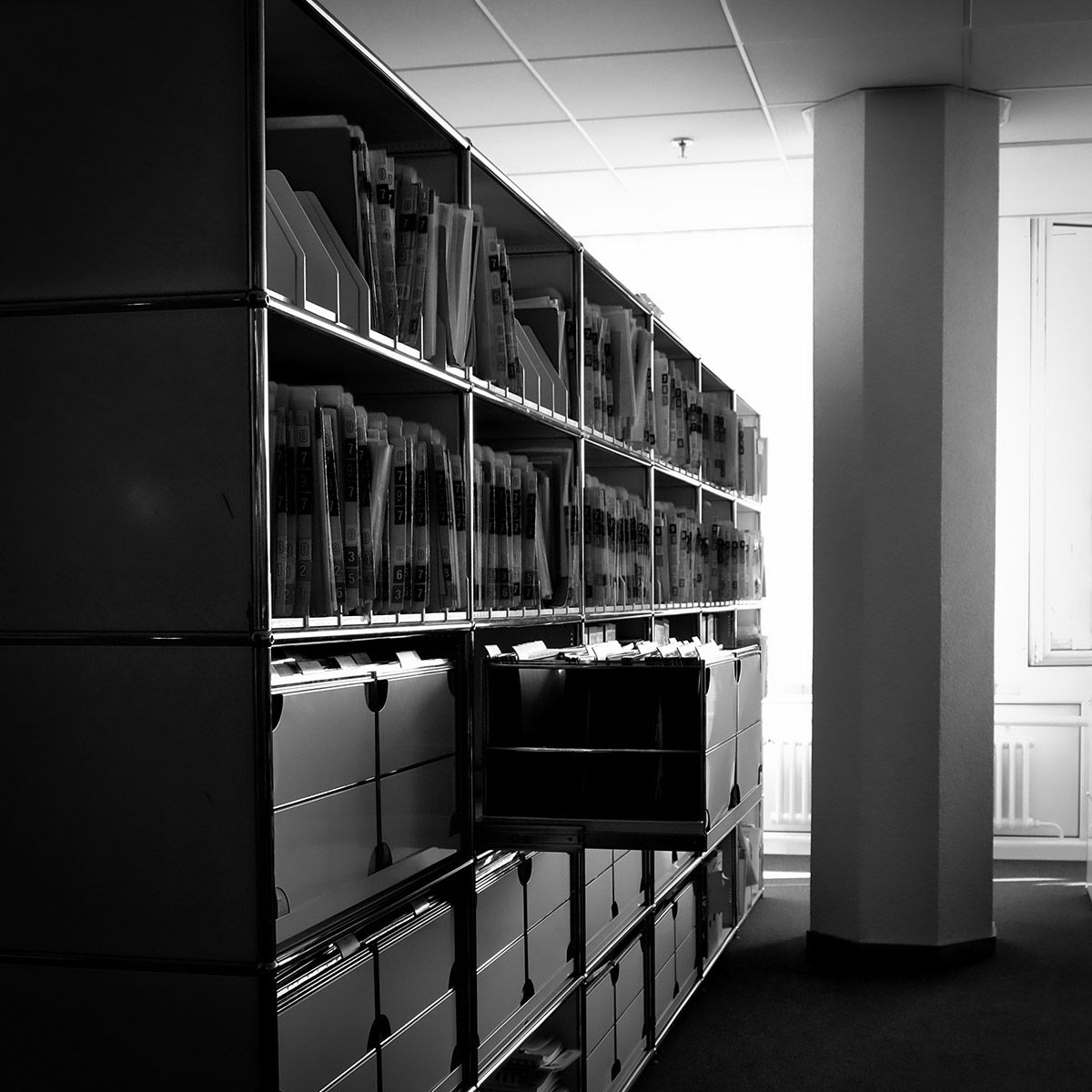 Des étagères remplies de dossiers dans une pièce avec un pilier et une fenêtre lumineuse.