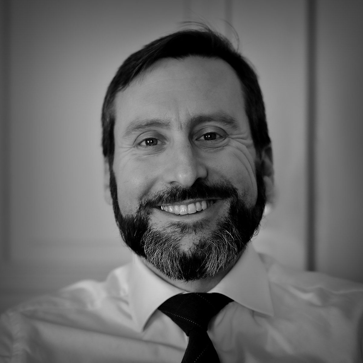 Un homme barbu, portant une chemise blanche et une cravate, affiche un large sourire sur une photo en gros plan, en noir et blanc.