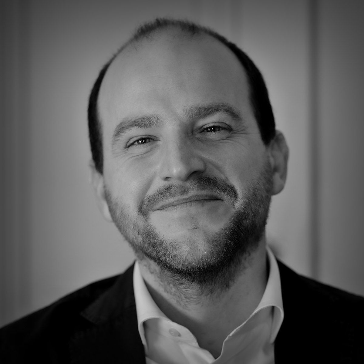 Homme souriant en veste de costume sombre, chemise blanche et barbe courte, à l'intérieur.