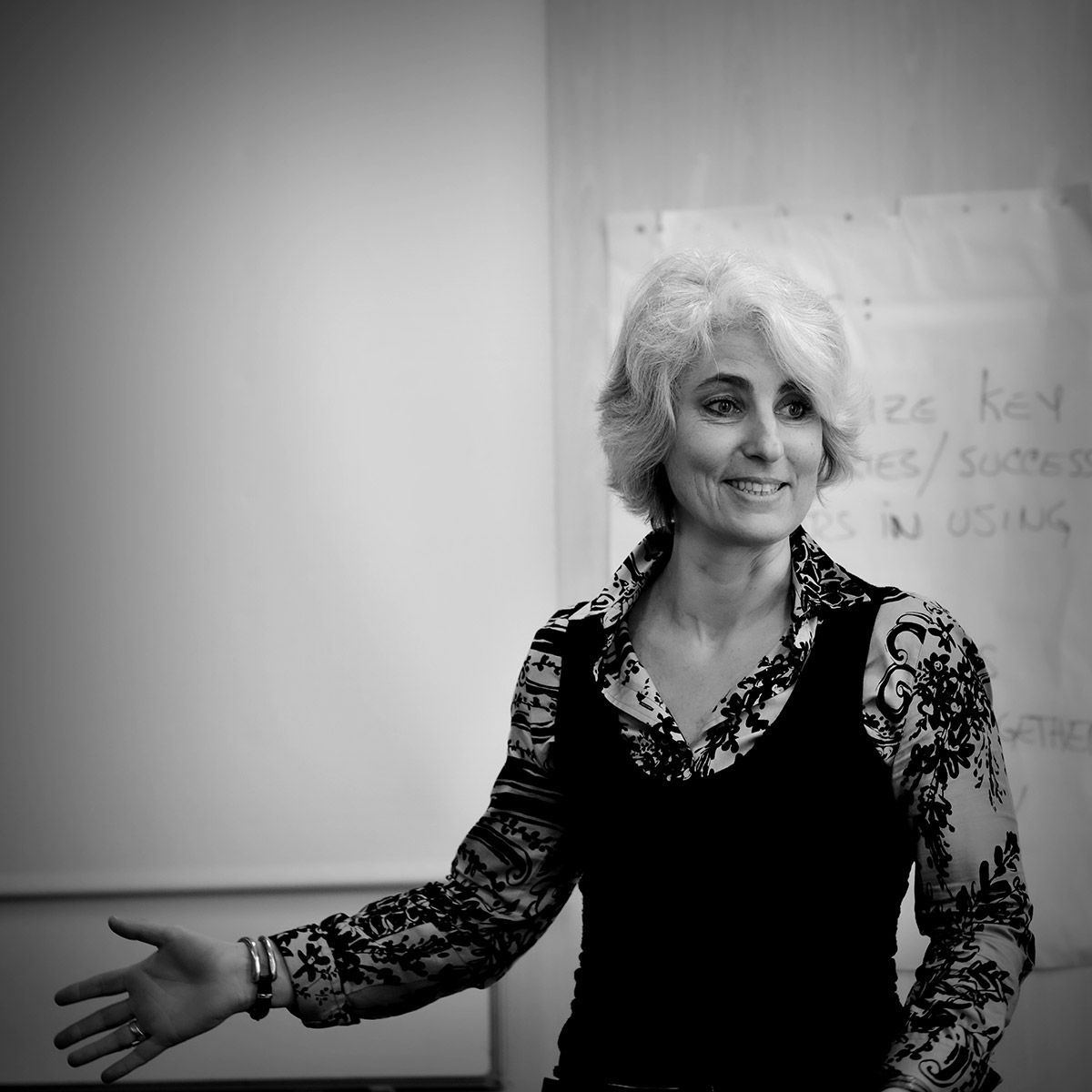 Une femme fait des gestes en souriant devant un tableau blanc. Elle porte une chemise et un gilet à fleurs.