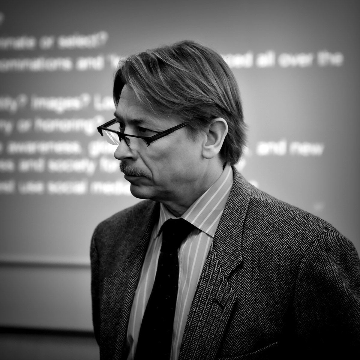 Un homme portant des lunettes, une cravate et un blazer, regarde sur le côté devant un écran de projection.