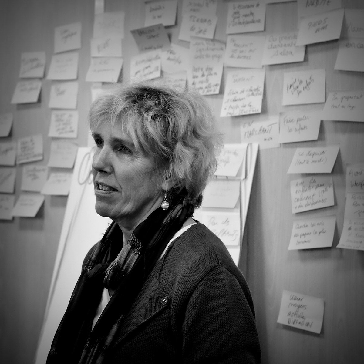 Une femme, debout devant un mur de post-it, parle. Elle porte un cardigan et une écharpe, et regarde vers sa gauche.