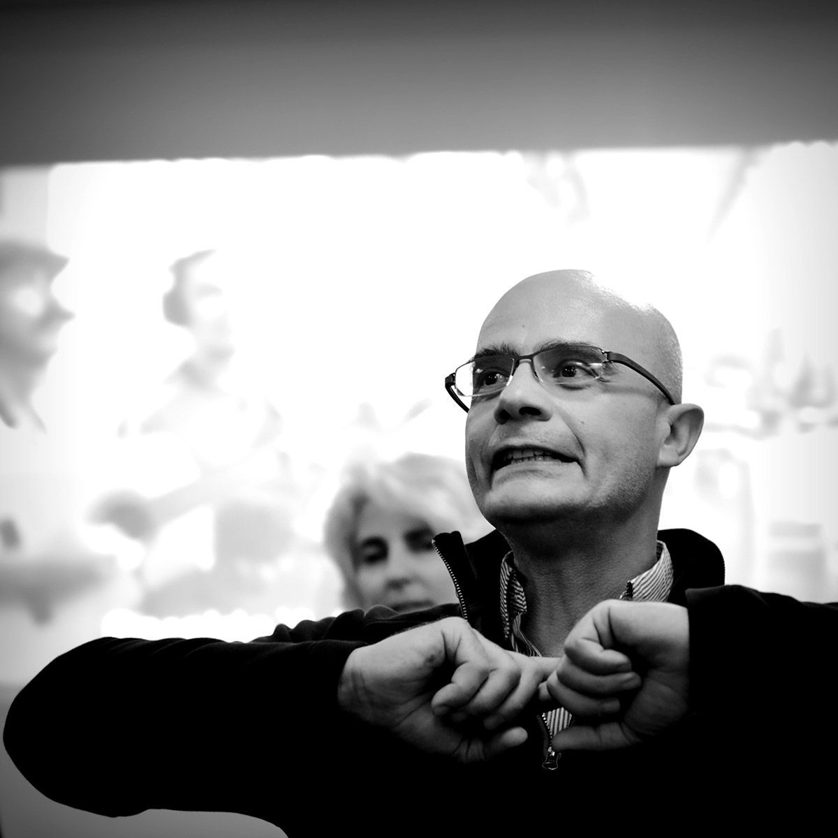 Un homme à lunettes gesticule avec enthousiasme à l'intérieur, une autre personne se devine derrière lui.